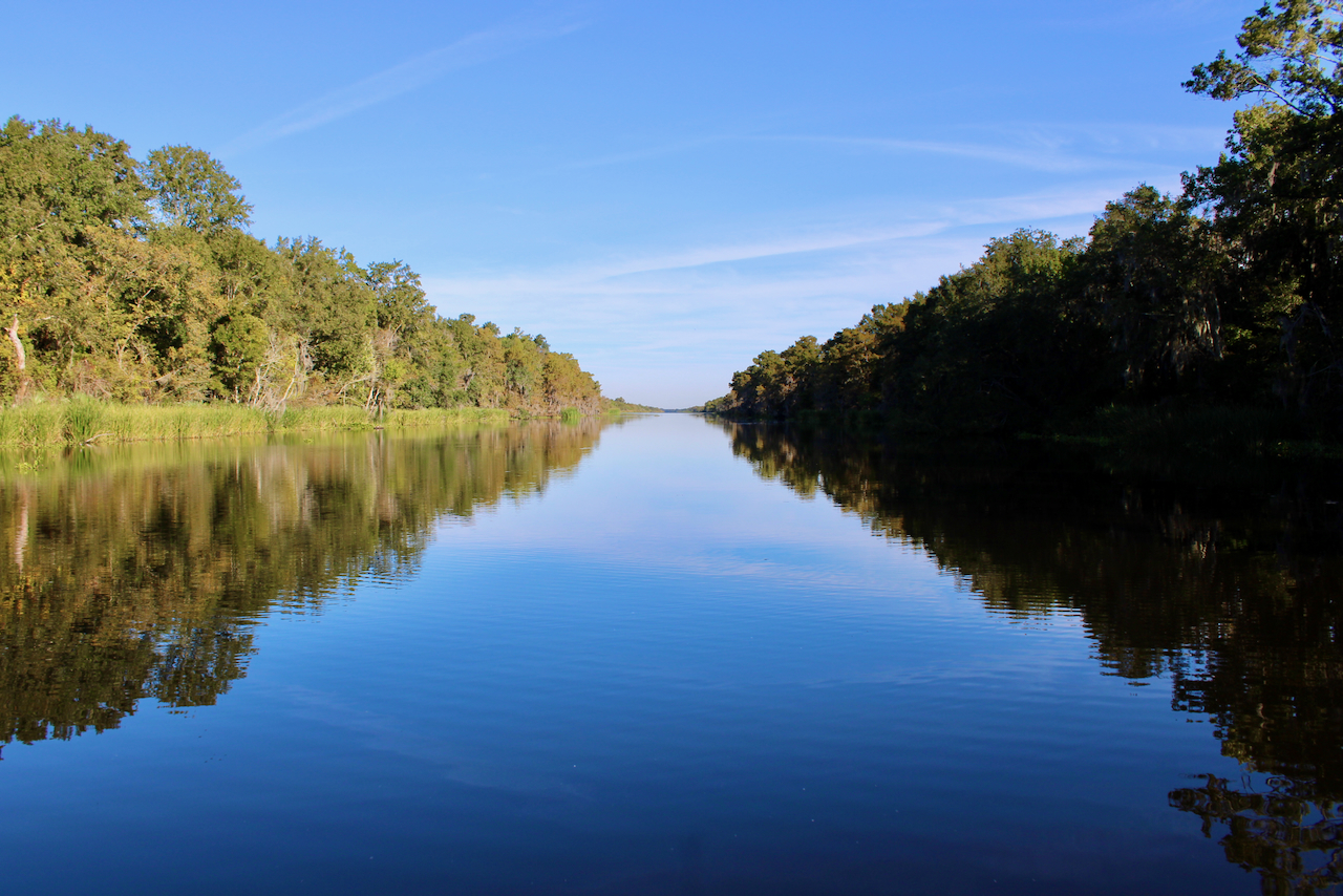 Photograph of a calm river reflecting lush trees under a clear blue sky.