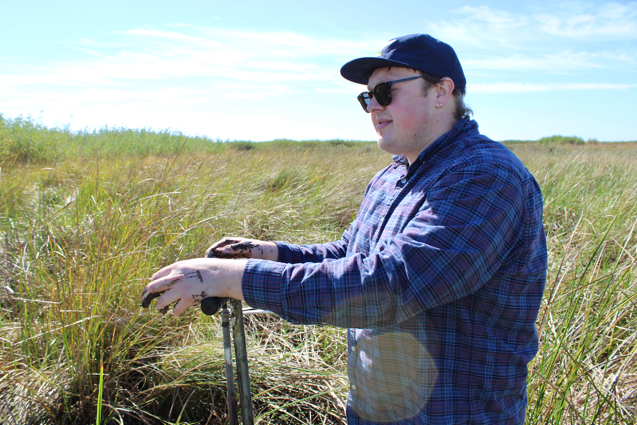 Photograph of a man in a blue plaid shirt and cap standing in a grassy field.