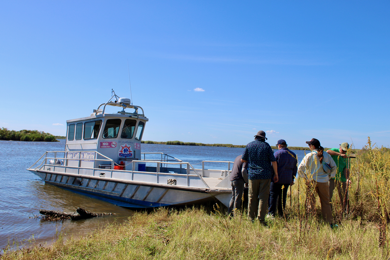 Photograph of a large, white research boat docked by a grassy riverbank with researchers nearby.