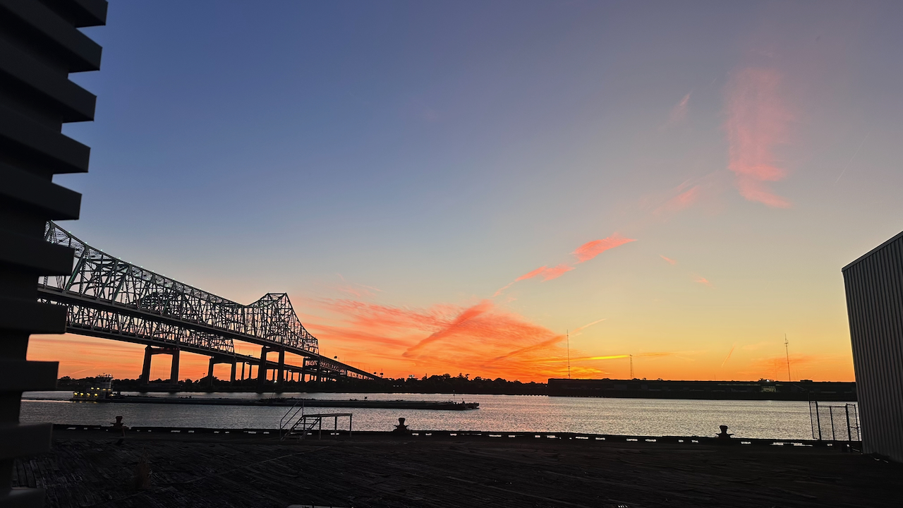 Photograph of a vibrant sunset over a bridge spanning a river, framed by city buildings.