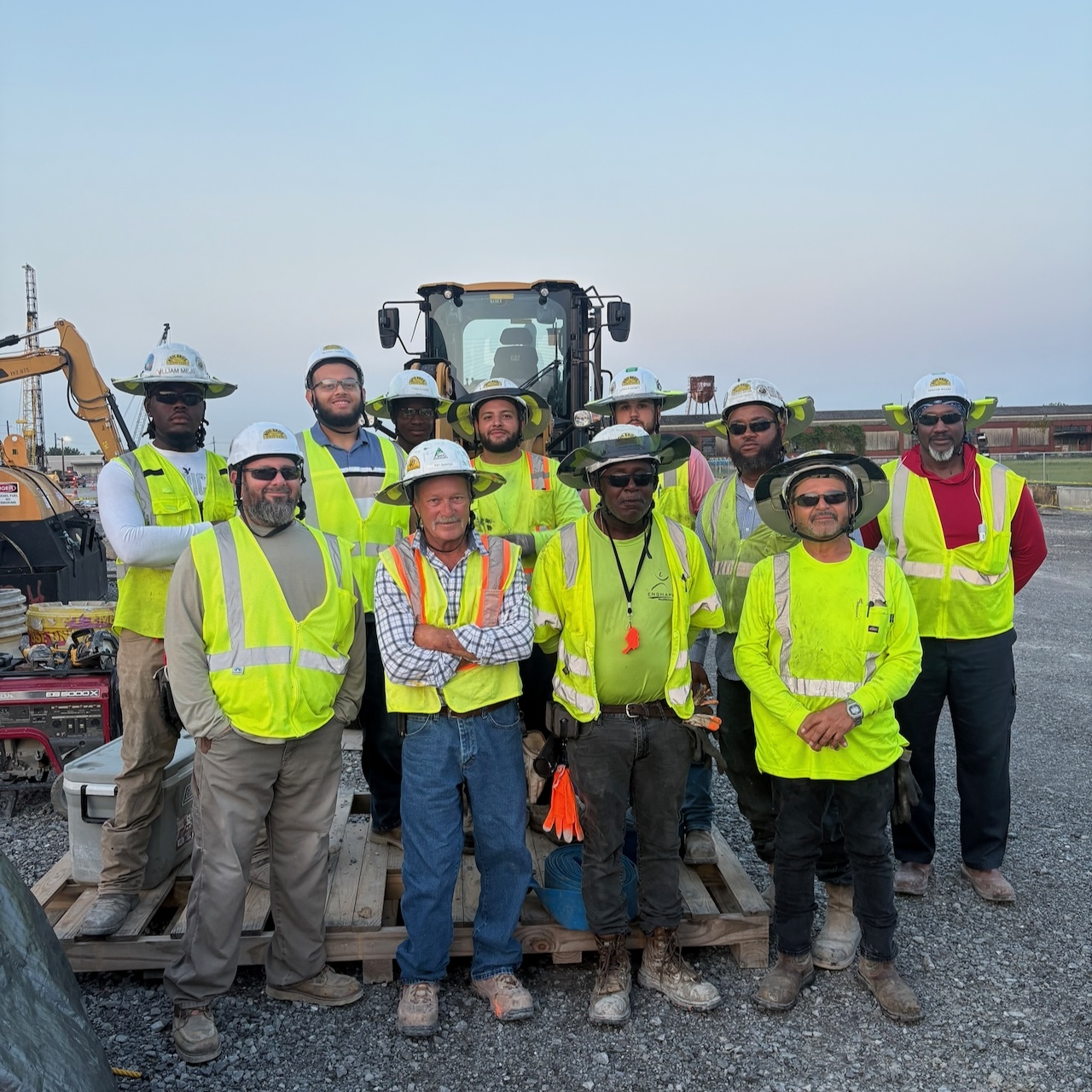 Photograph of ten construction workers in high-vis vests and hard hats standing before a tractor.