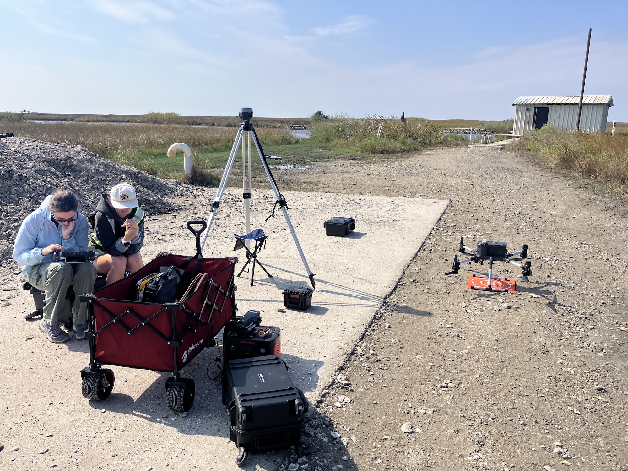 Two people sitting on the ground next to a red wheel barrow and a drone