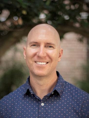 Portrait of a smiling bald man in a blue patterned shirt outdoors.