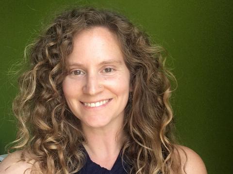 Photograph of a woman with long curly brown hair smiling against a green background.