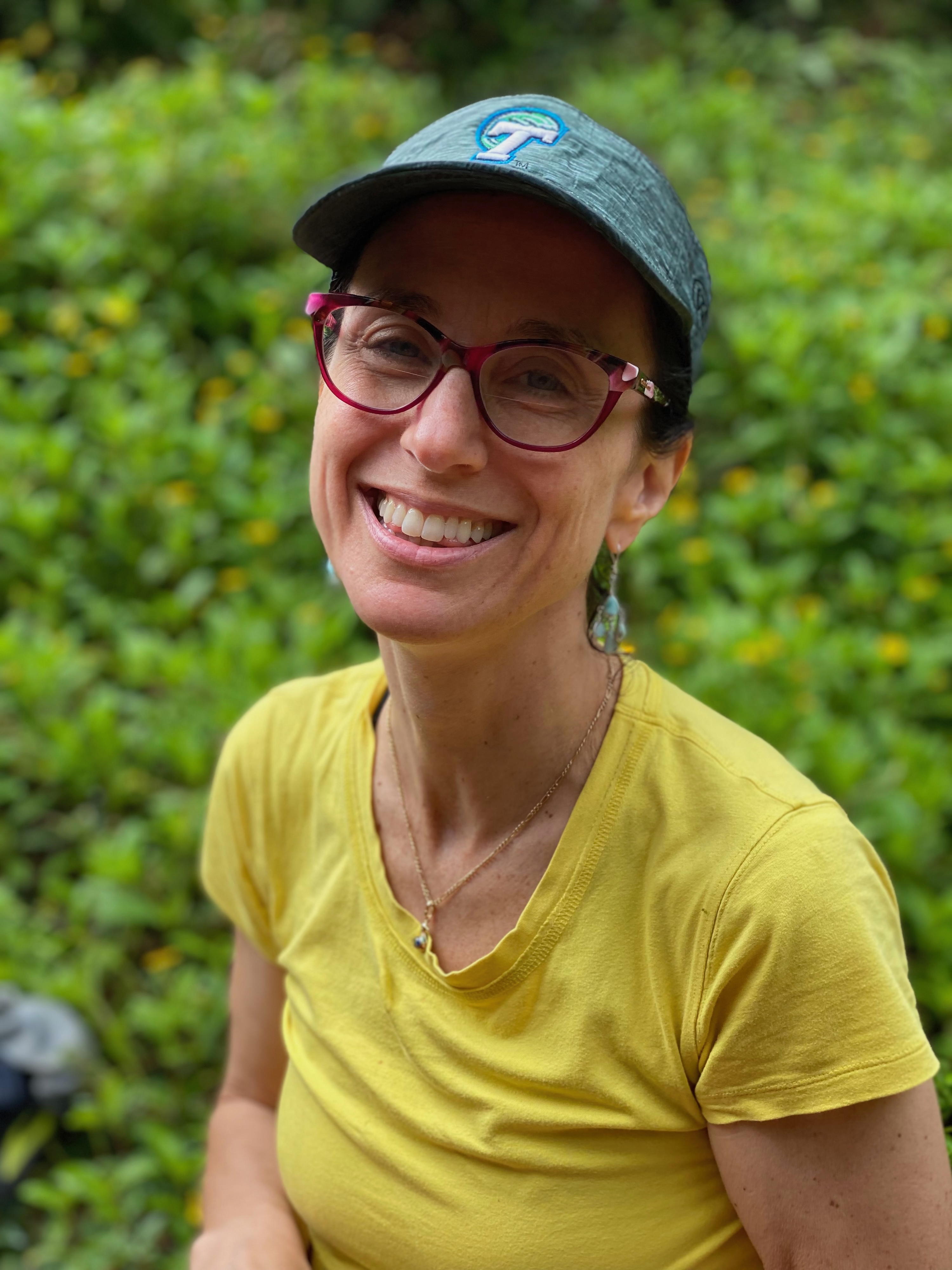 Photograph of a smiling woman in a yellow shirt and cap amidst green foliage.