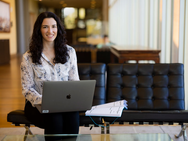 Photograph of a smiling woman with dark hair sitting on a black couch, holding a laptop.