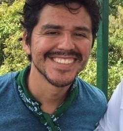 Close-up photo of a smiling man with dark hair, a mustache, and a blue shirt outdoors.