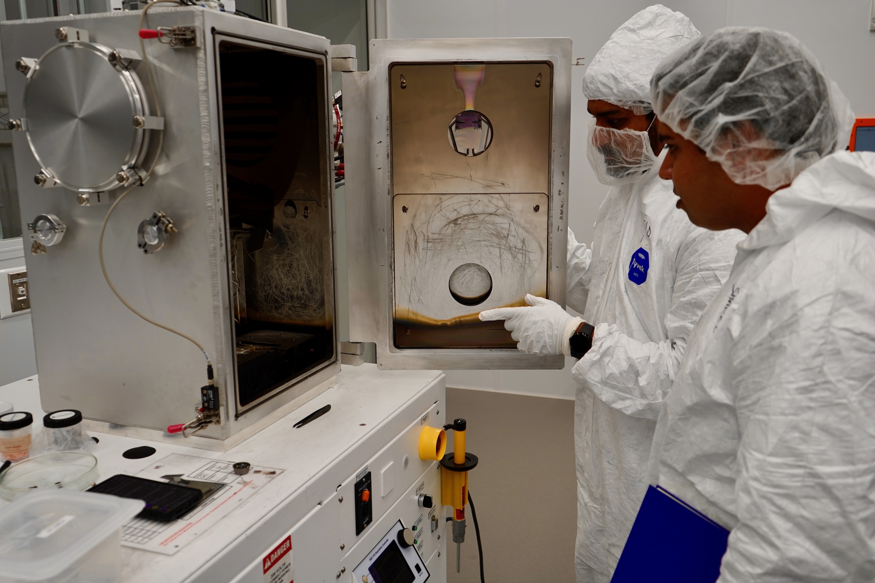 Two researchers in cleanroom suits examine an open laboratory chamber.
