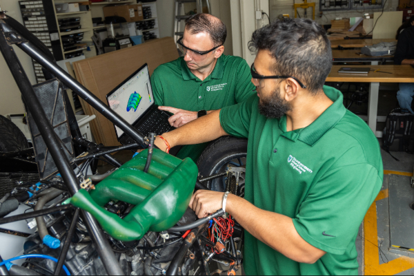 Photograph of two men in green polo shirts working on a car in a garage.