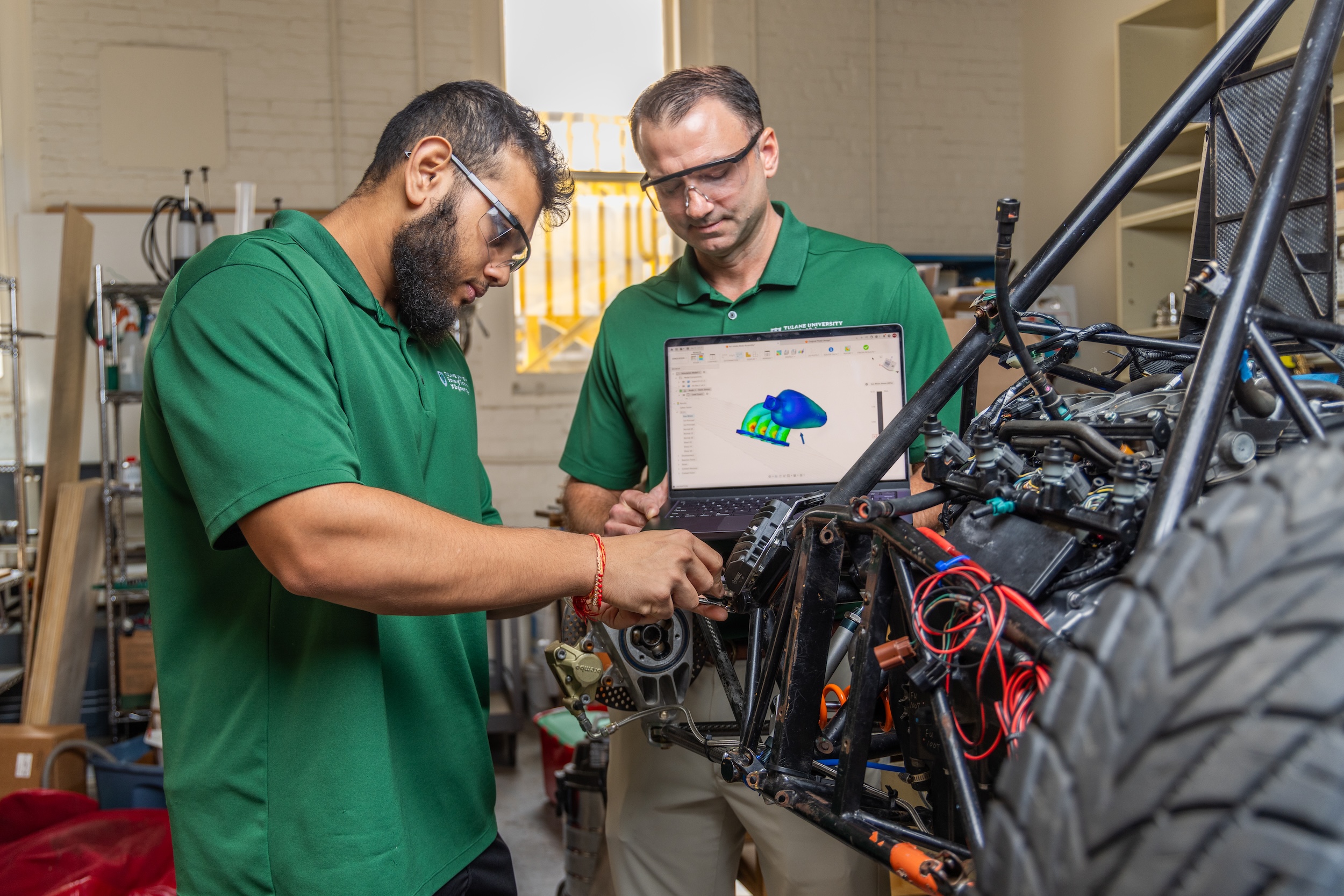 Photograph of two male engineers in green shirts building a race car frame in a workshop.