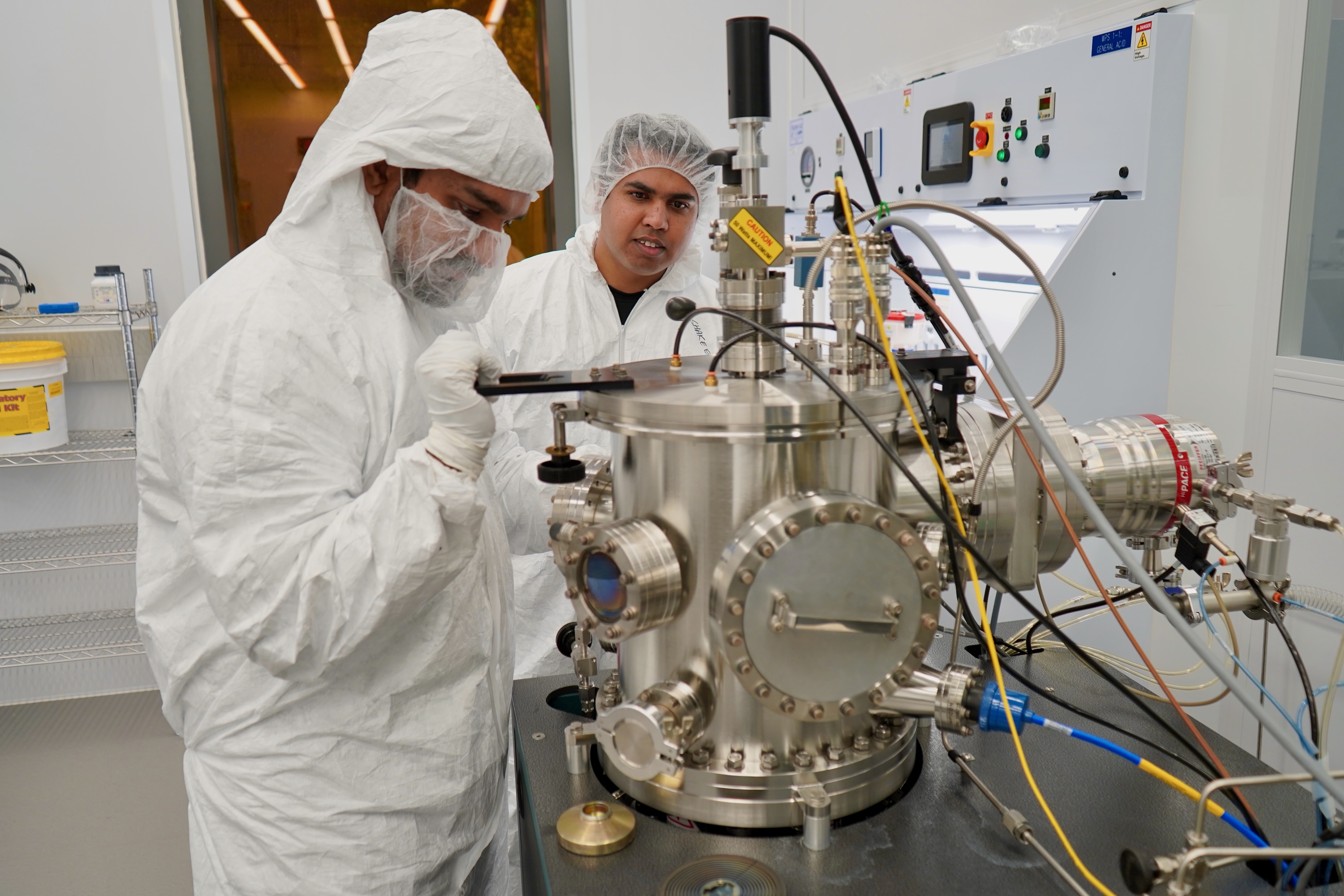 Two researchers in cleanroom gear operate a complex scientific chamber.