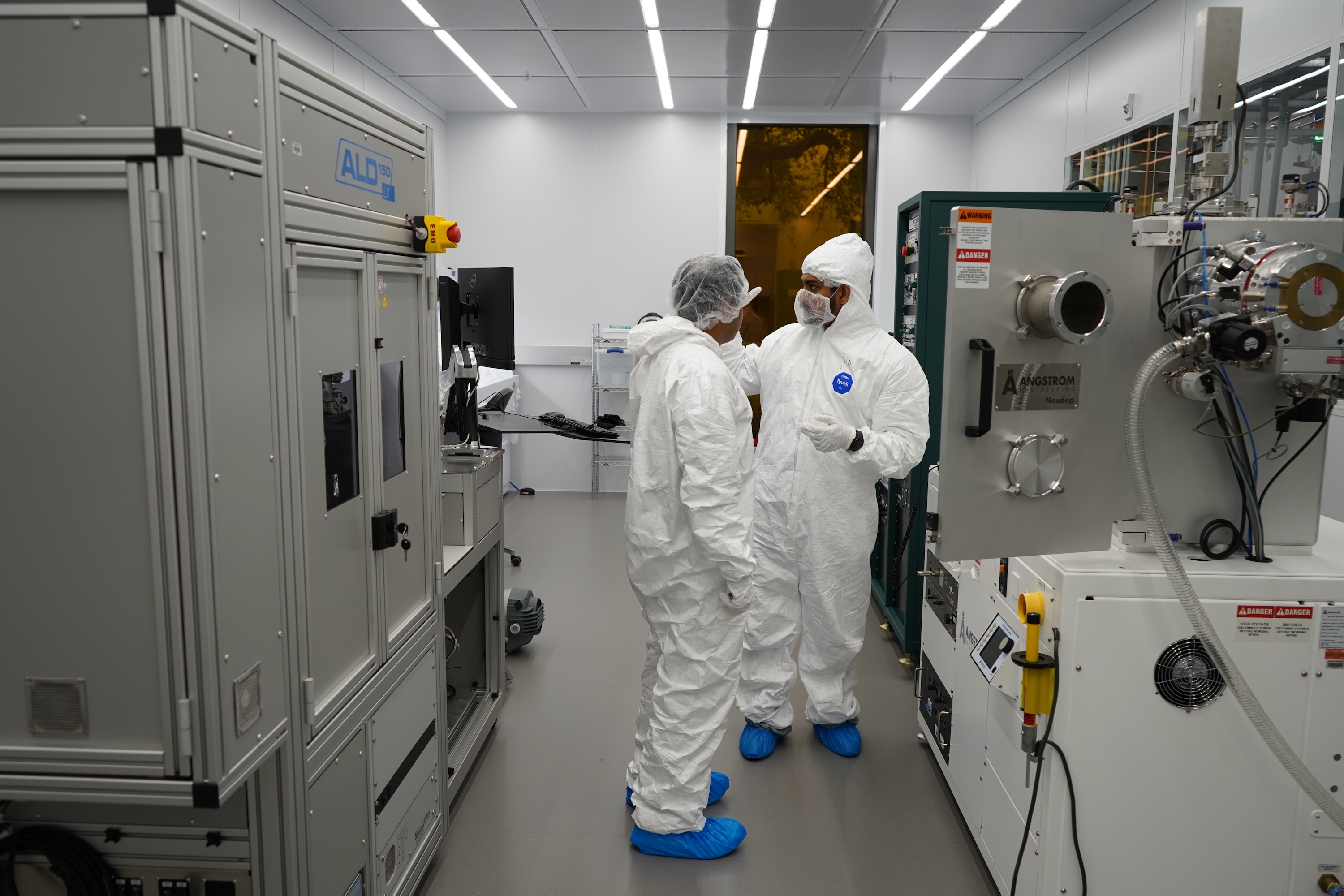Two technicians in white cleanroom suits and blue booties work in a lab with heavy machinery.
