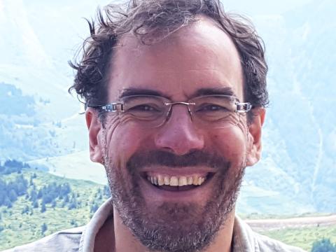 Headshot photograph of a smiling man with glasses, curly brown hair, and a beard outdoors.