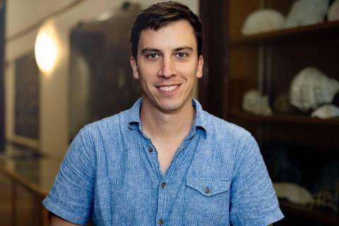 Photograph of a smiling man in a blue button-up shirt standing indoors.