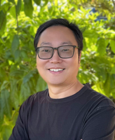 Photograph of an East Asian man wearing glasses smiling against a green leafy background.