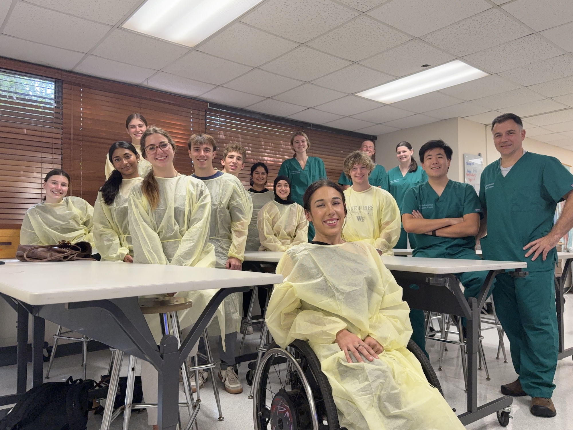 Group photo of students and instructors in a medical classroom wearing scrubs and gowns.