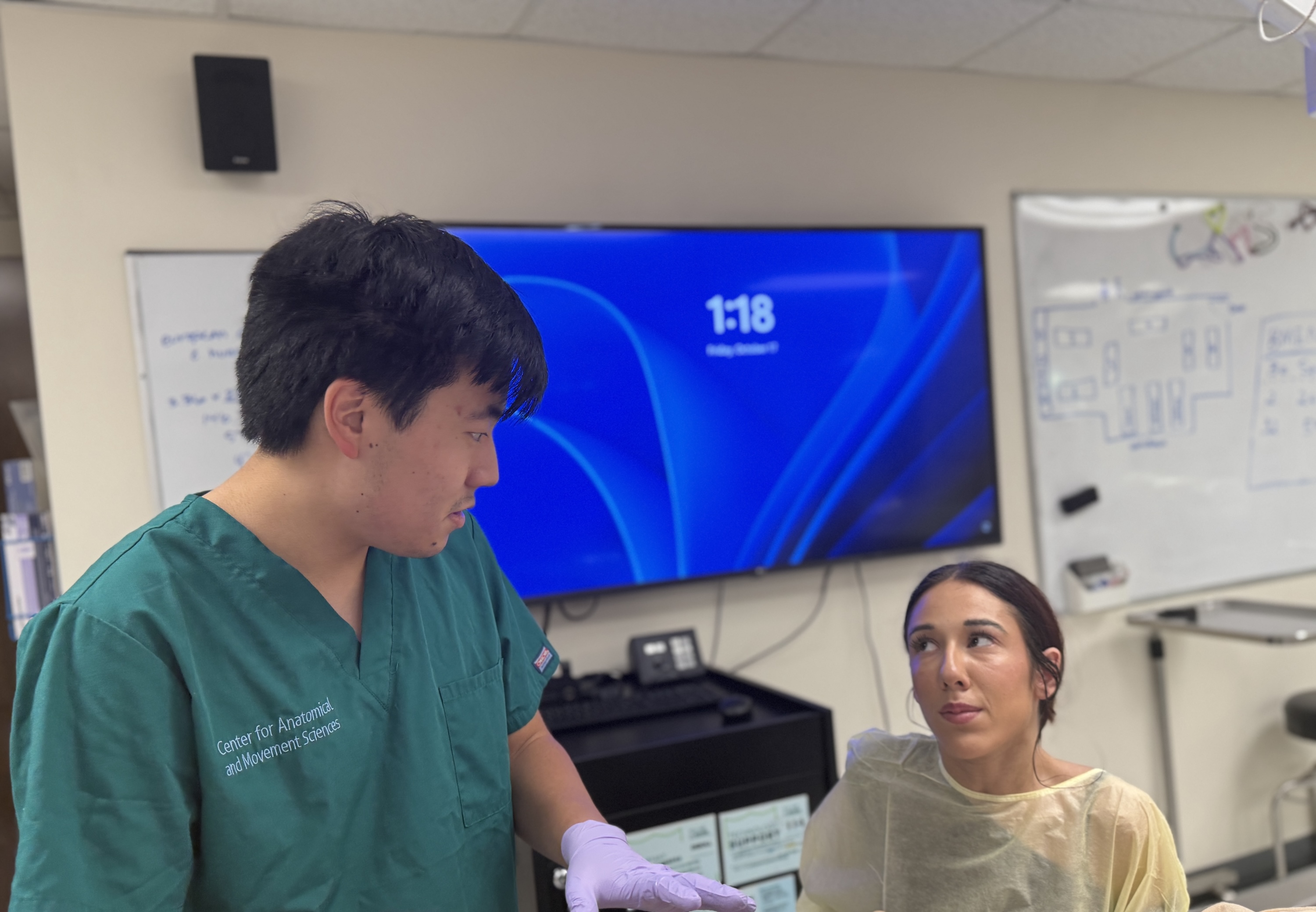 A male clinician in green scrubs talks to a patient wearing a yellow gown in a medical room.