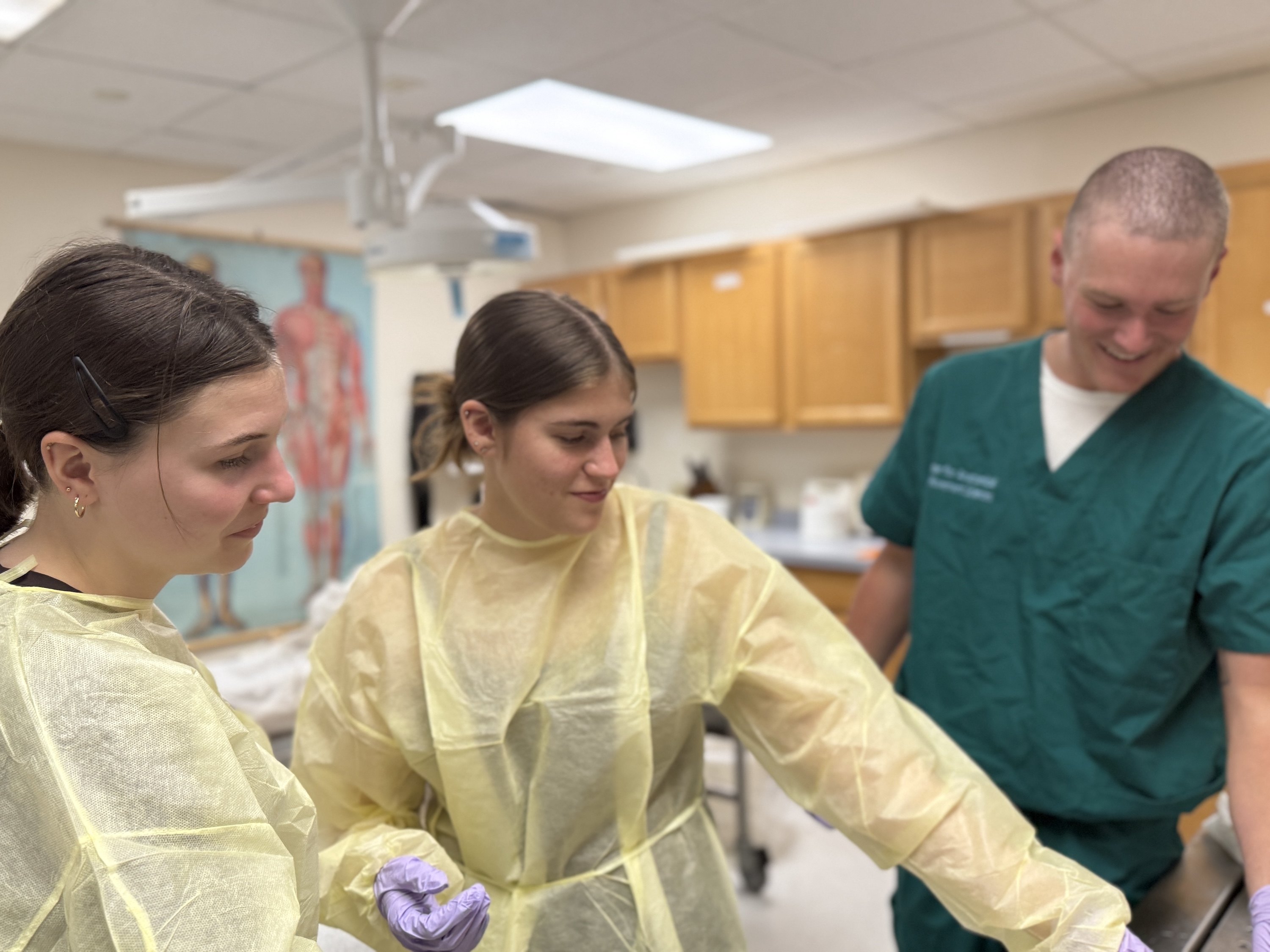 Photo of three students in scrubs and gowns smiling together in a clinical lab setting.