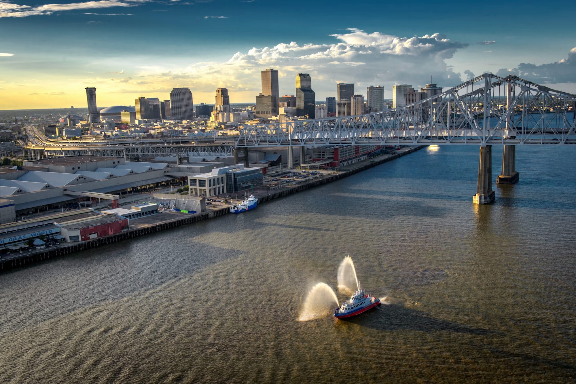 Photograph of a fireboat spraying water in a river near a city skyline and bridge at sunset.