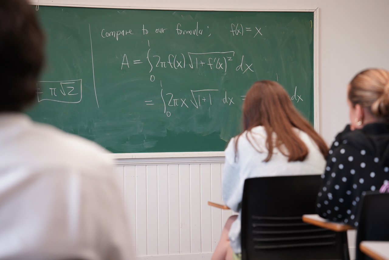 Students seated in a classroom facing a green chalkboard with calculus formulas.