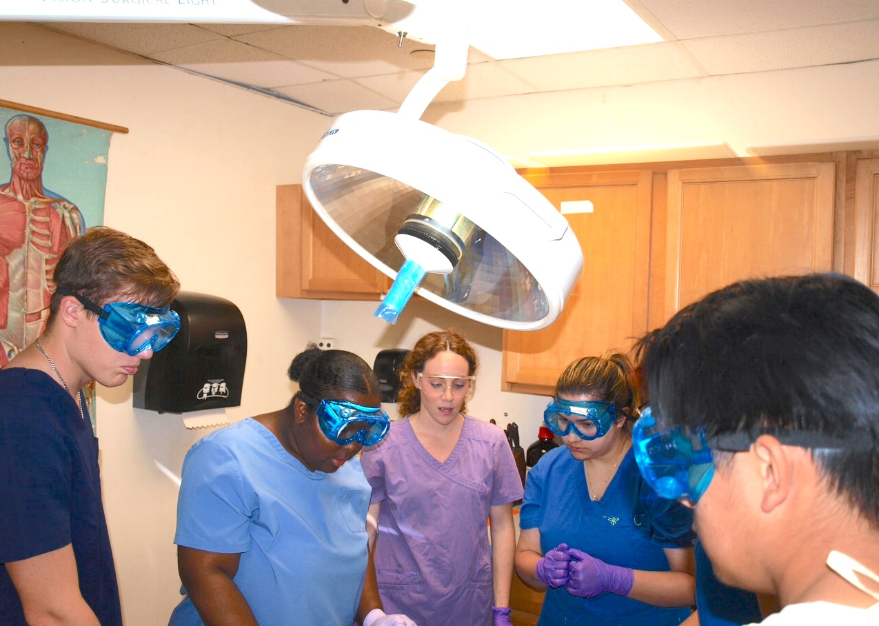 Students in scrubs and safety goggles gather around a lab table beneath a surgical light.