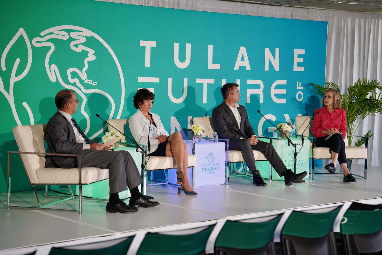 Photograph of a panel discussion with four people seated on armchairs in front of a teal backdrop.