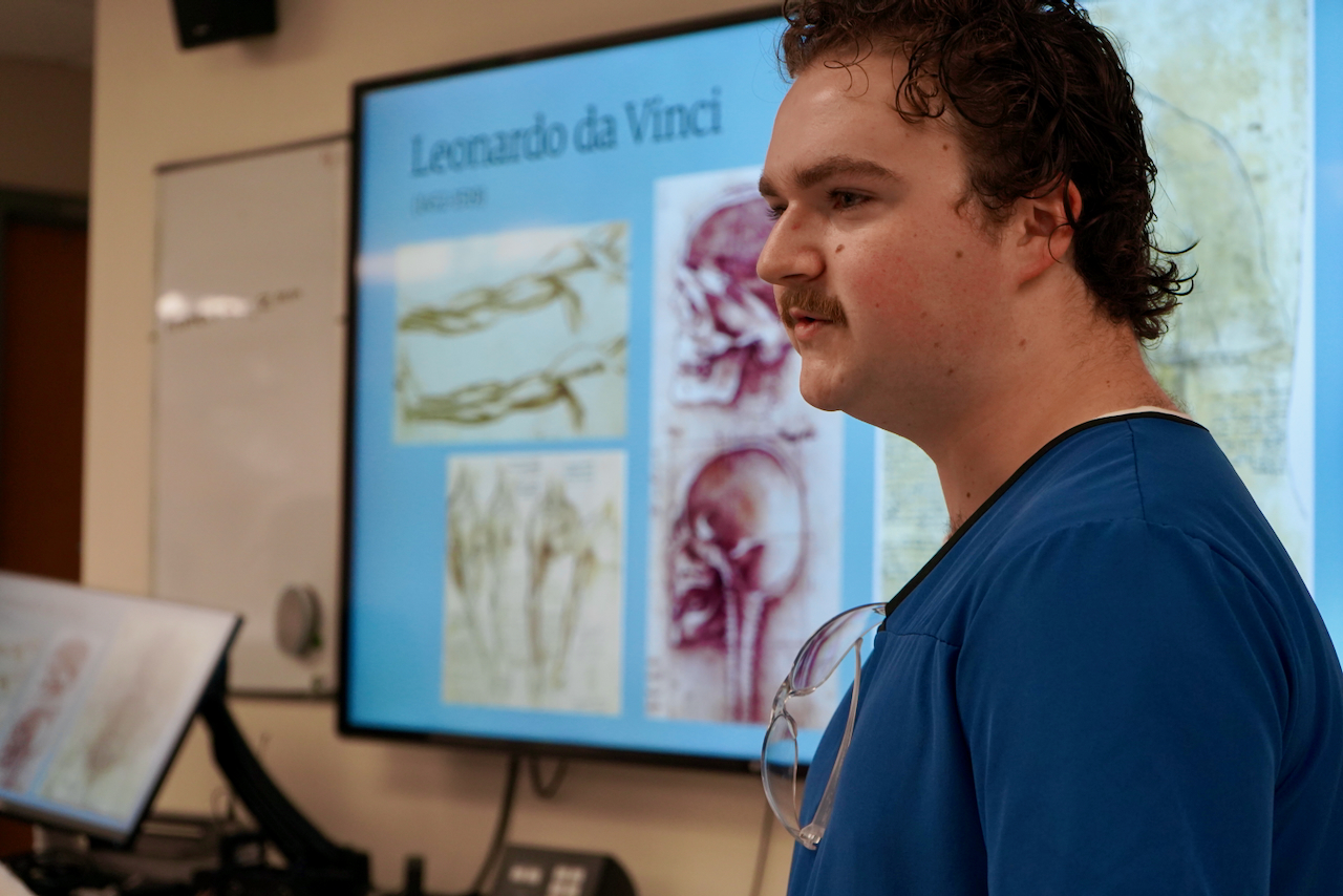 A man with a mustache in a blue shirt standing in front of a presentation screen about Leonardo da Vinci.