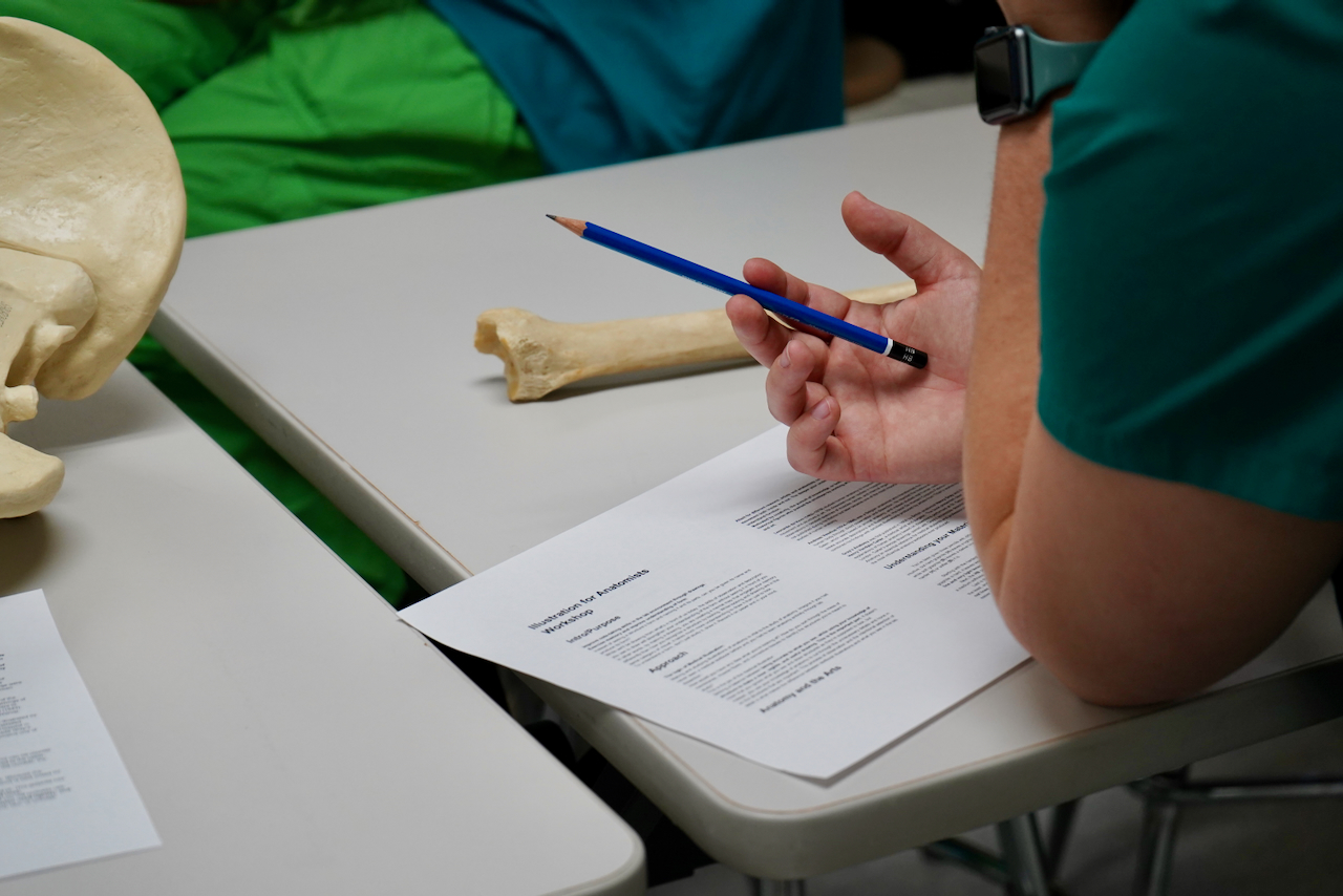 A person in a green shirt sits at a desk, holding a blue pencil above a paper.
