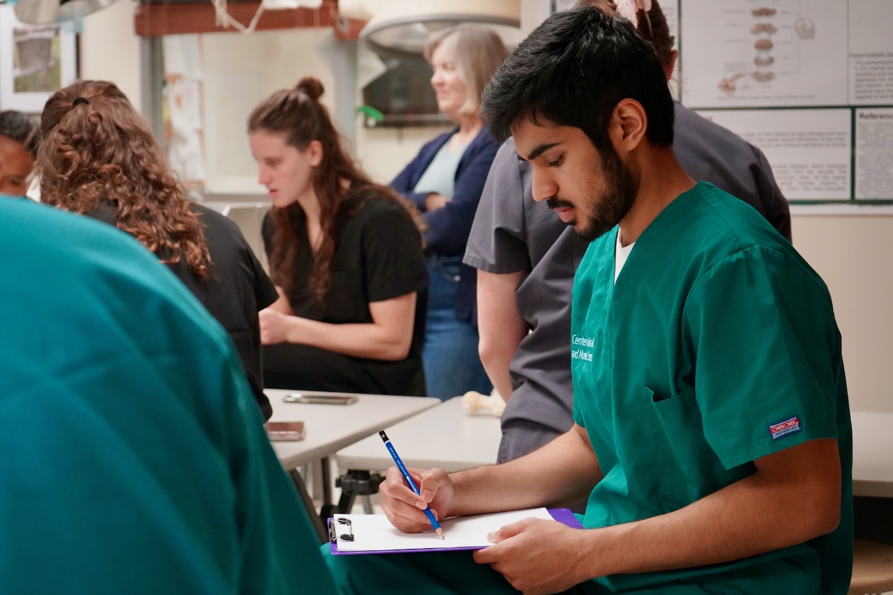 Photograph of a man in green scrubs writing on a clipboard in a classroom.