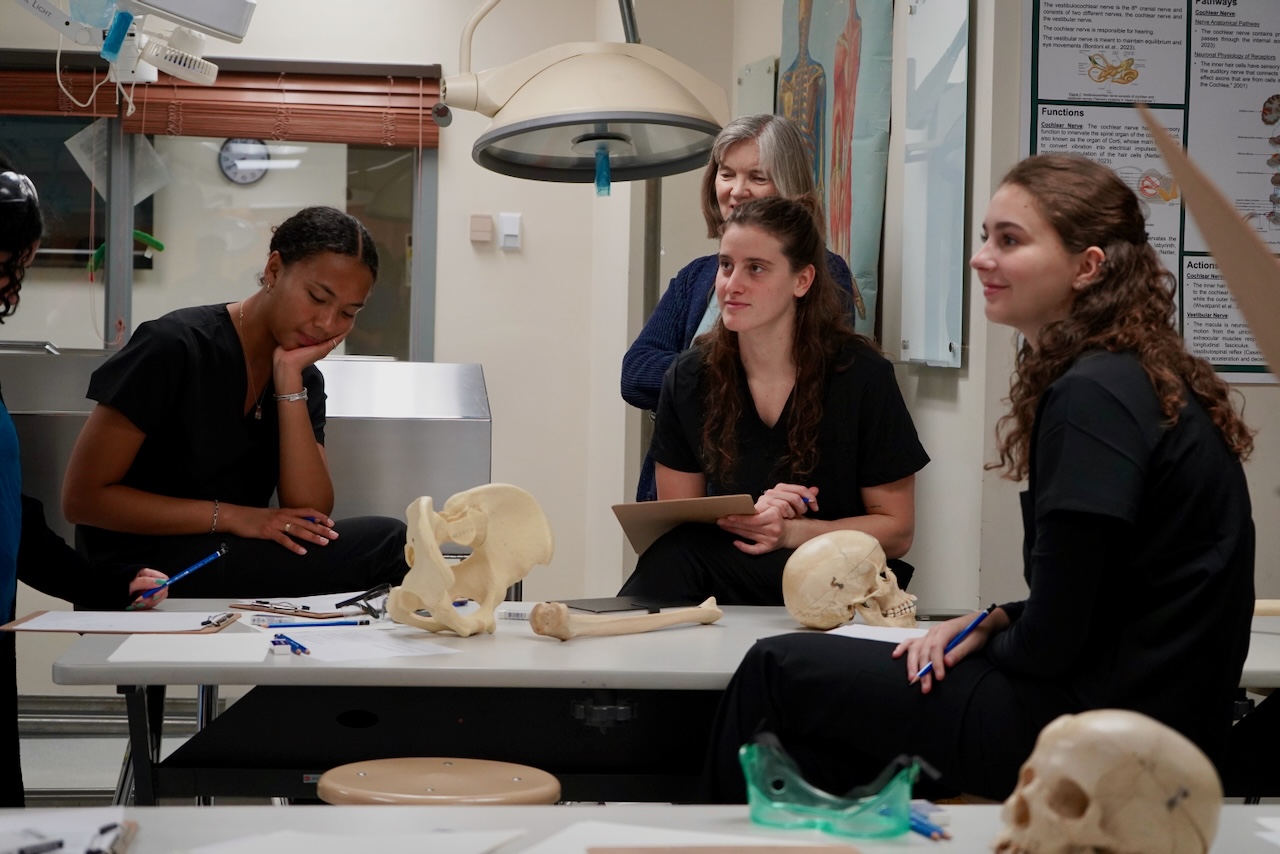 A photograph of female students studying anatomy with bones on a table in a classroom.