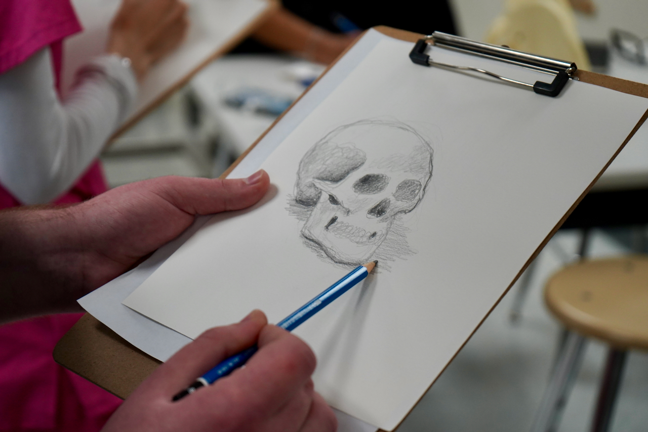 Photograph of a person drawing a skull on a clipboard with a blue pencil.