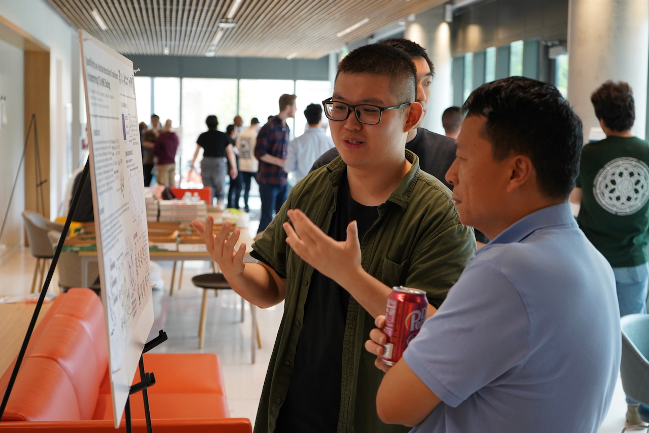Photograph of a man in a green shirt explaining a research poster to another man.