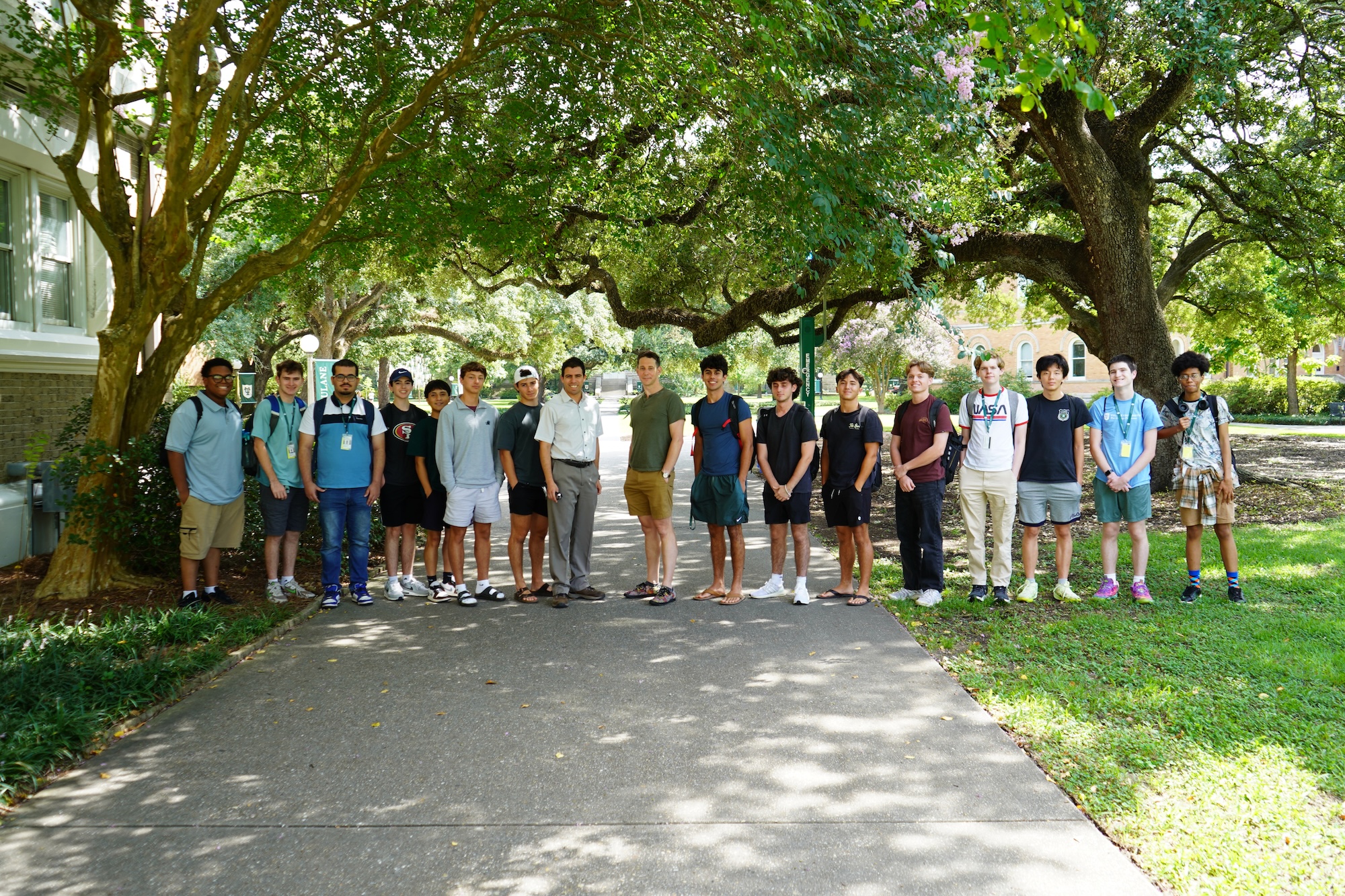 Photograph of a large group of teenage boys standing on a paved walkway beneath large trees.