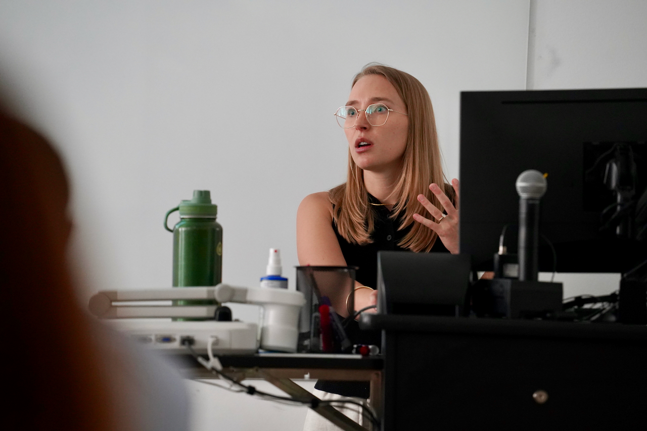 A blonde woman wearing glasses speaks at a desk with a microphone.