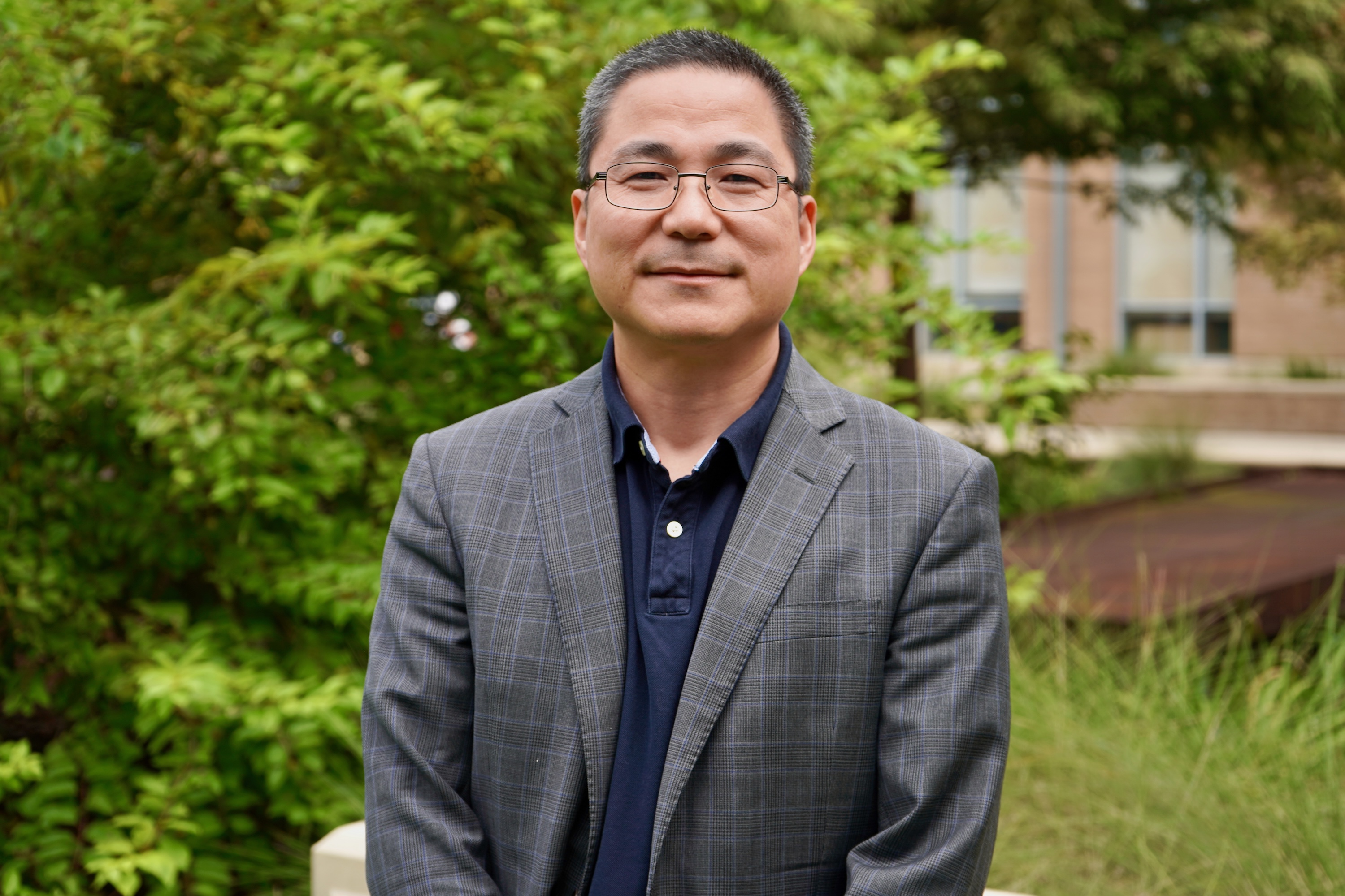 Photograph of a smiling Asian man wearing glasses and a grey plaid blazer outdoors.