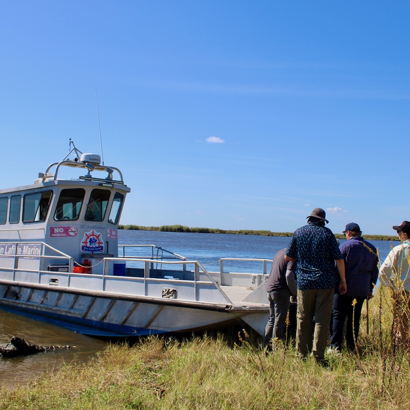 Photograph of four people standing by a boat docked on a grassy riverbank.
