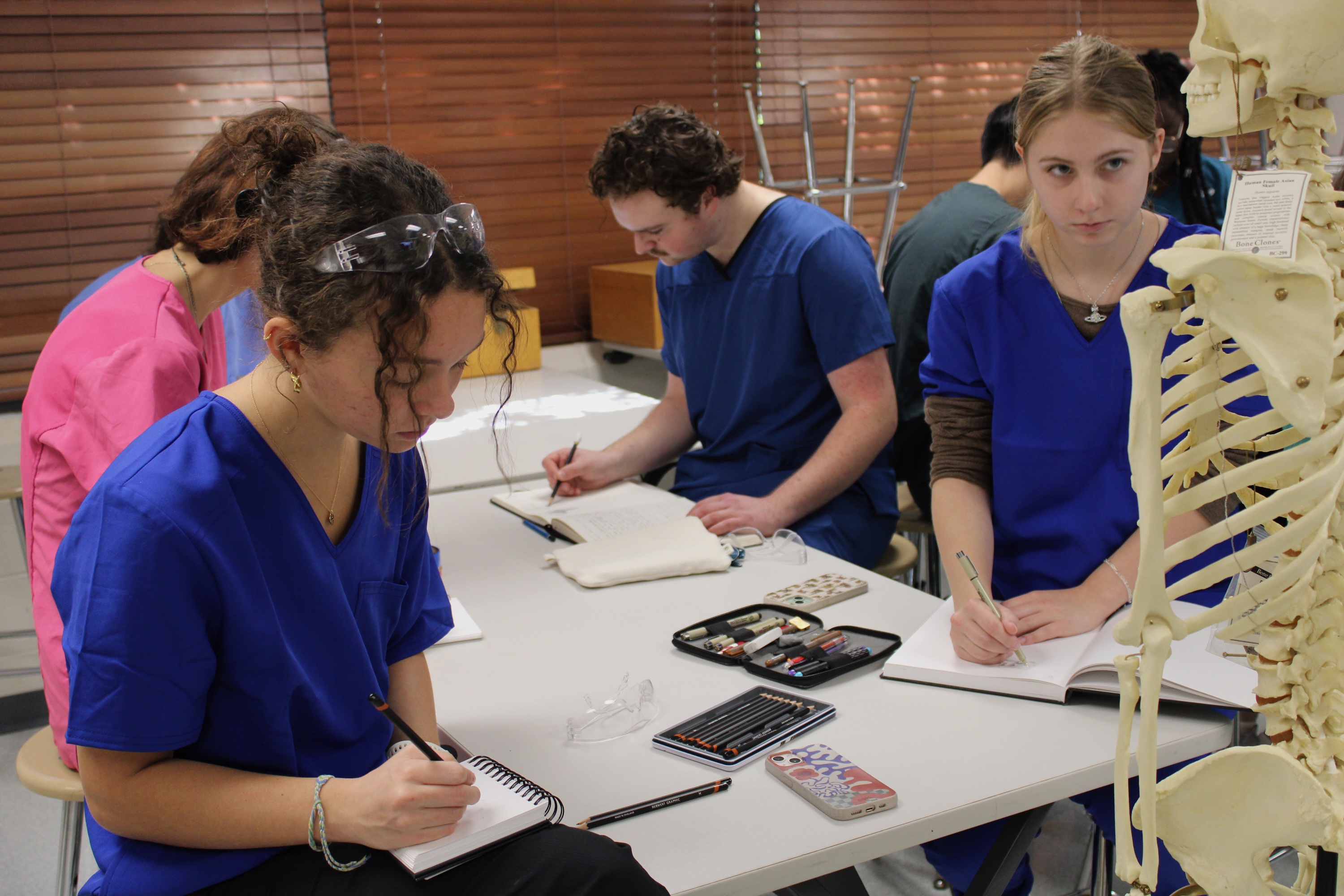 Photograph of students in blue and pink scrubs writing at a table with an anatomical model.
