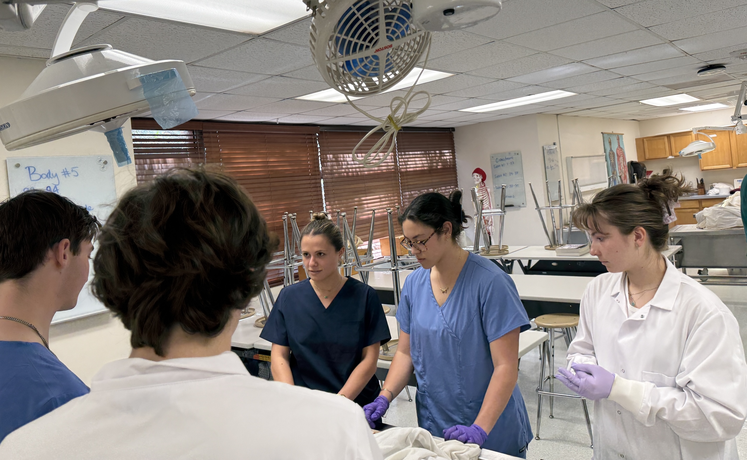 Students in medical scrubs gather around a lab table in a brightly lit classroom.