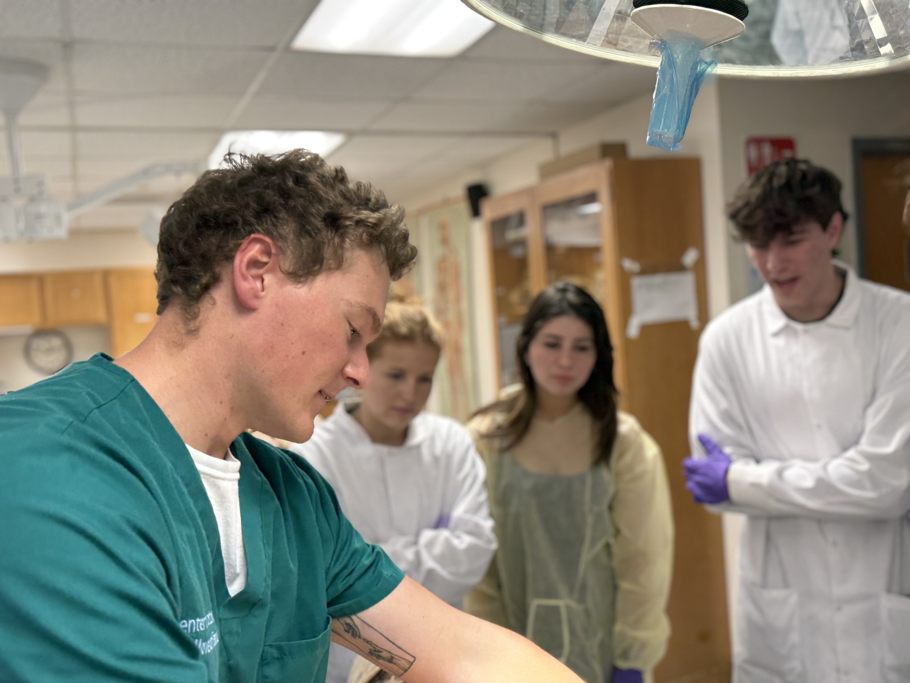 A photograph of a student in green scrubs examining a model while three peers watch intently.