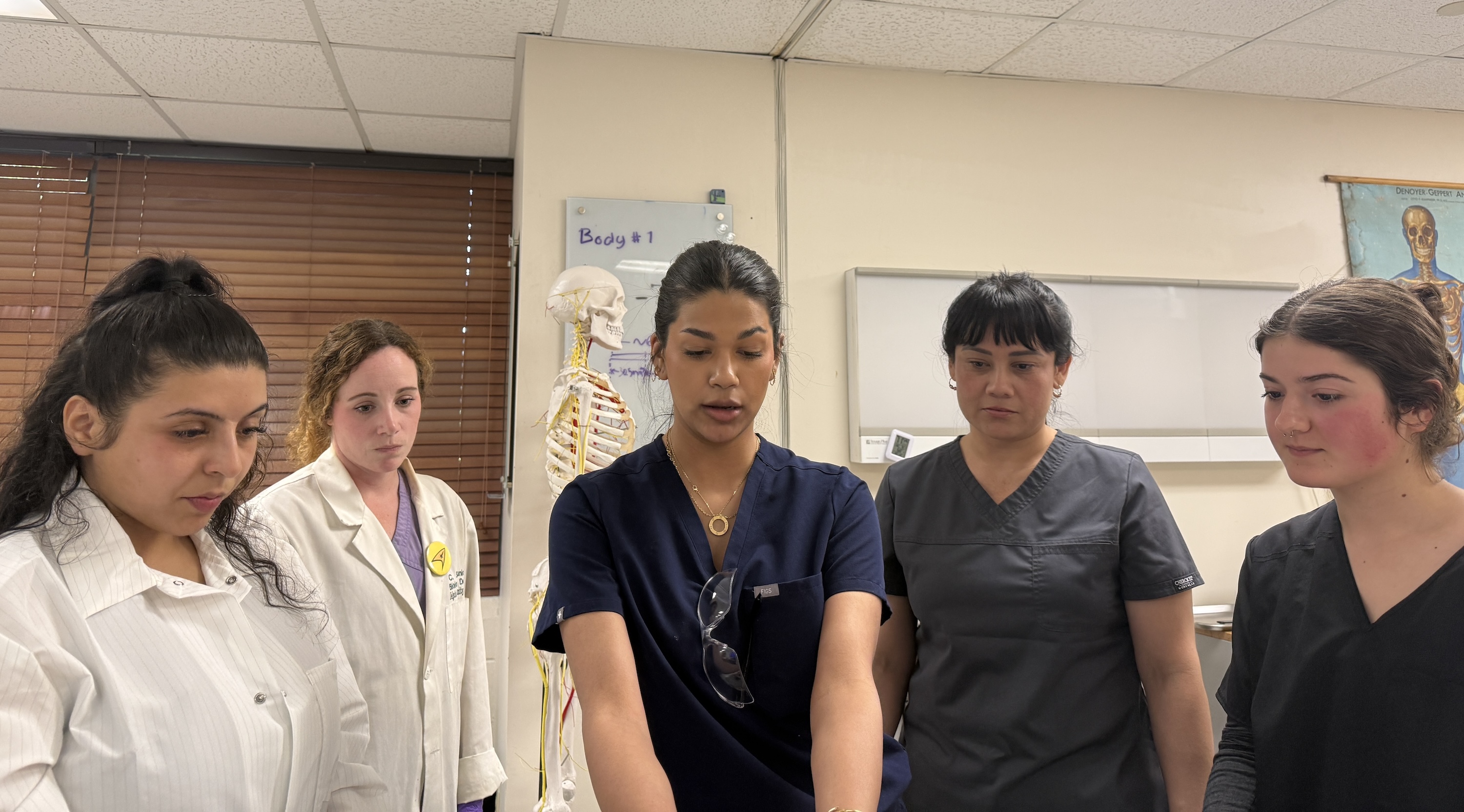 Five medical students in a classroom, with one speaking and the others listening intently.