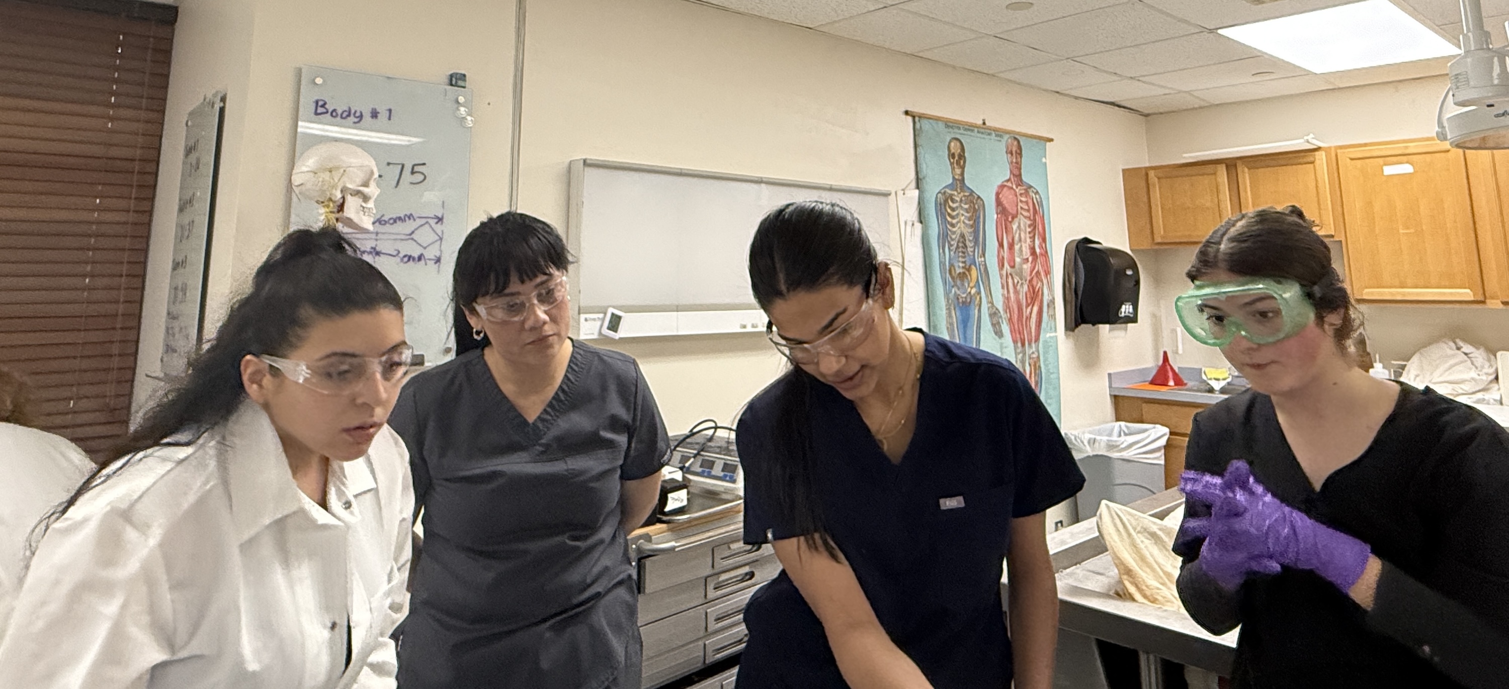 Four young women in medical scrubs gather around a table in a classroom.