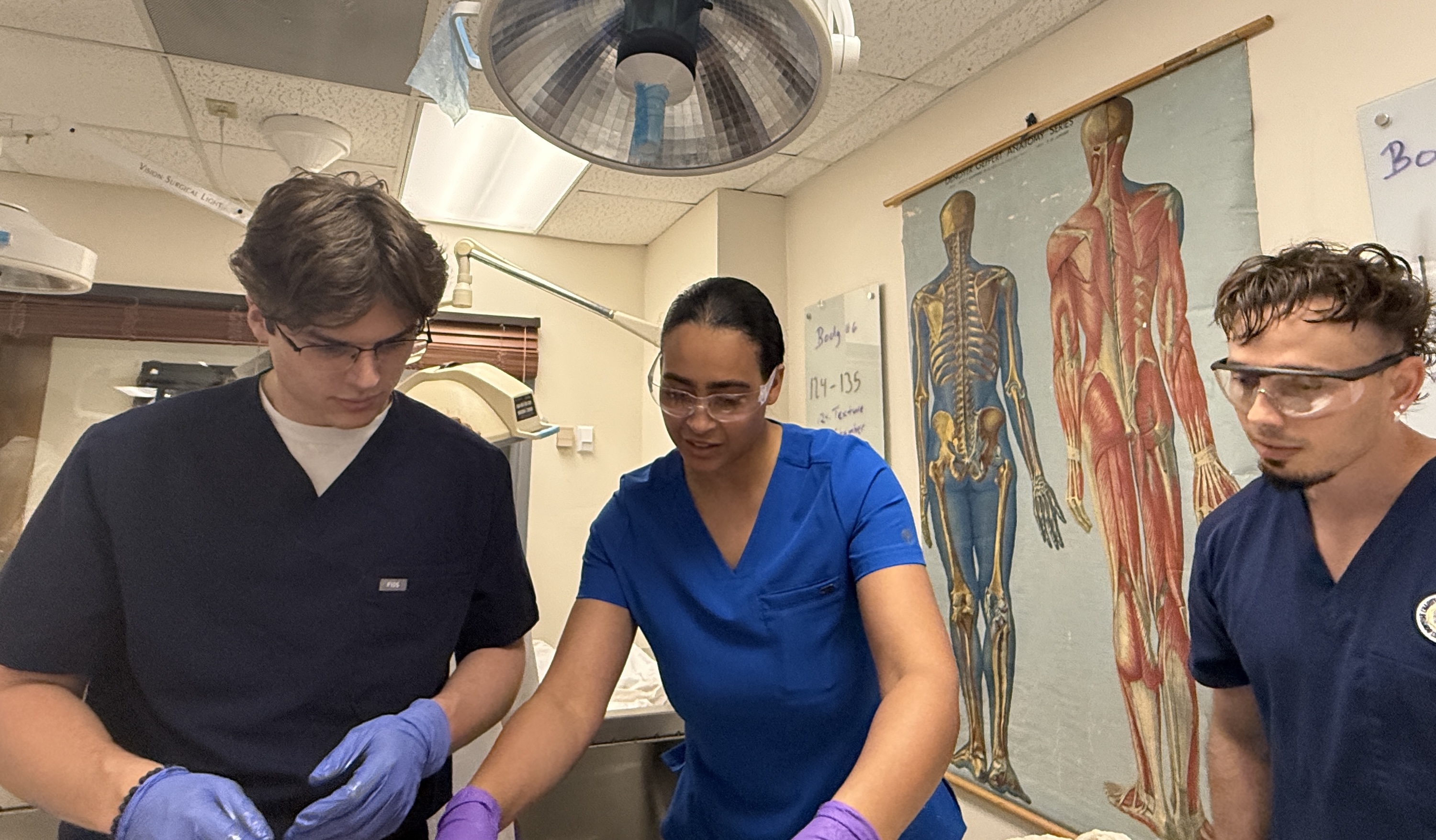 Photograph of three students in scrubs, focused on a task in a medical lab.