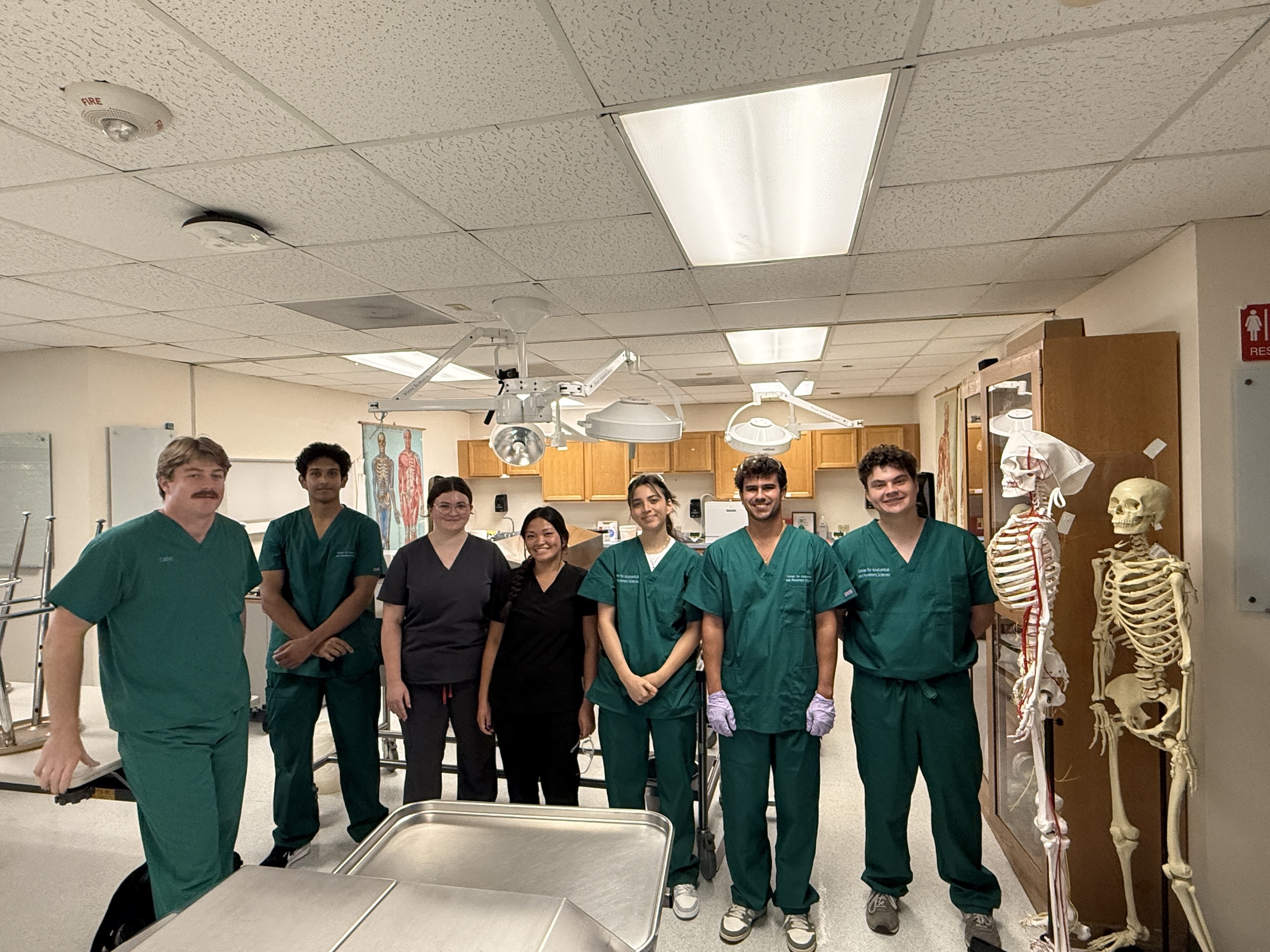 Photograph of seven medical students in scrubs posing in a lab with anatomical models.