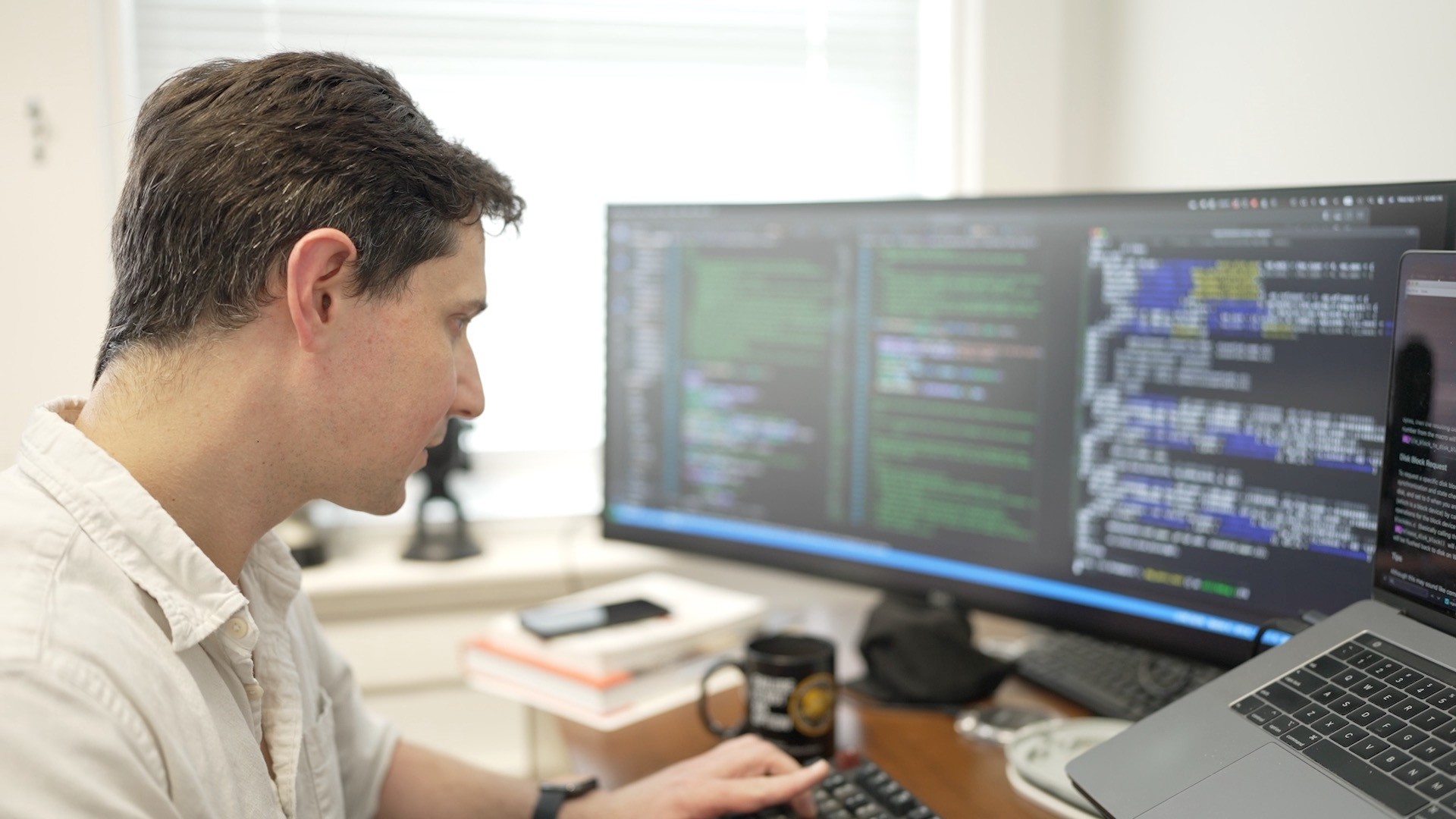 Photograph of a man coding in front of a wide monitor and laptop.