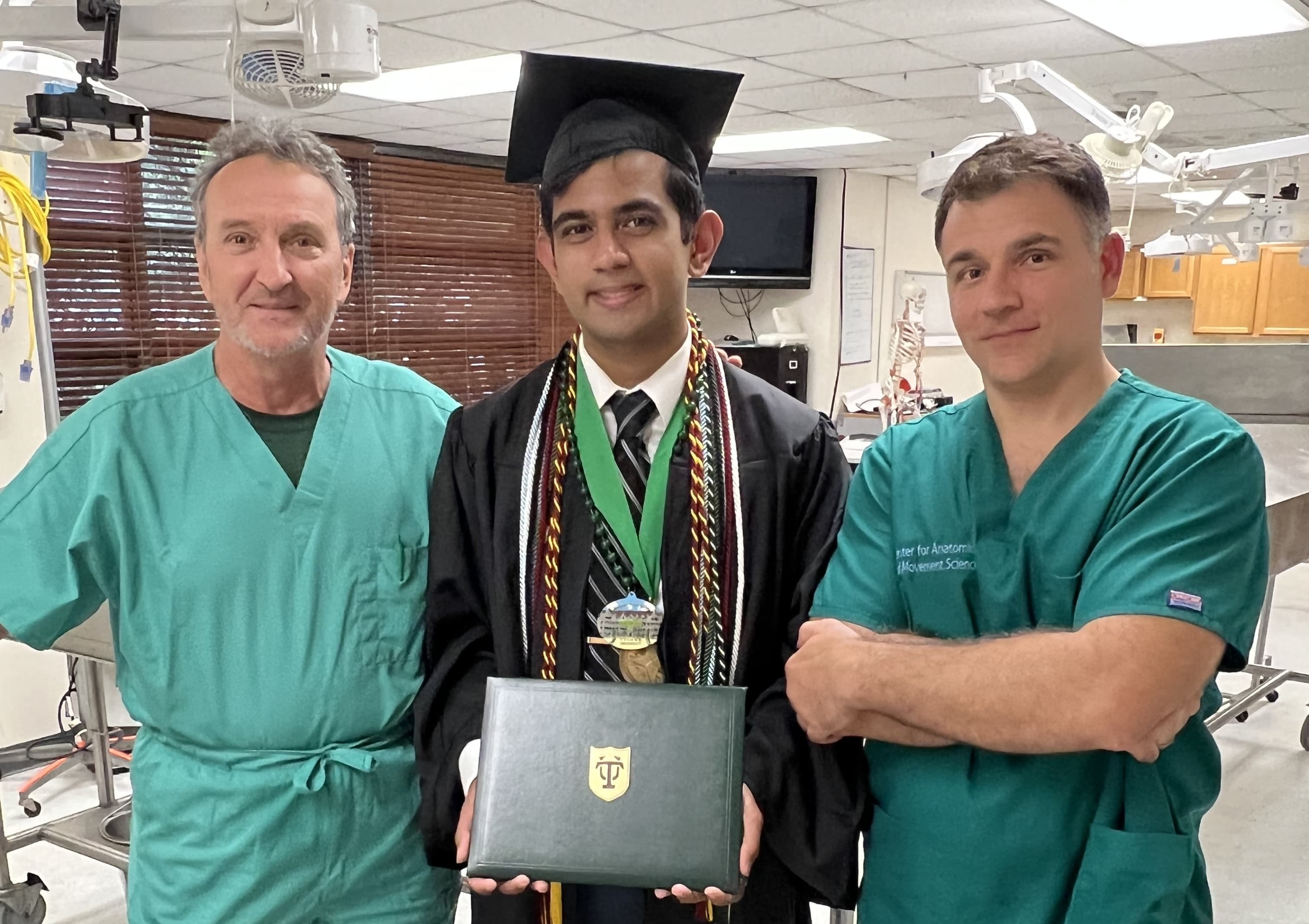 Photograph of a graduate in cap and gown holding a diploma, flanked by two men in scrubs.