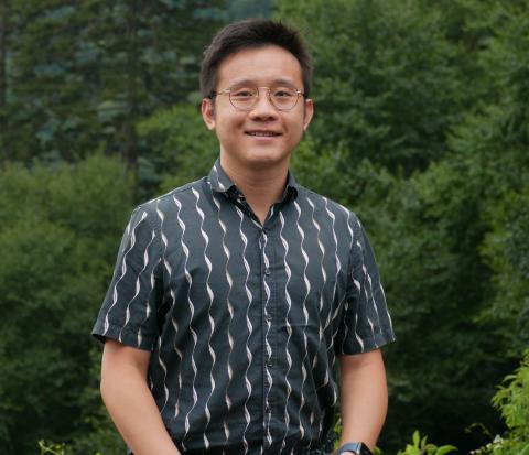 Photograph of a smiling Asian man wearing glasses and a patterned shirt outdoors.