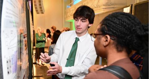 A student in a white shirt and green tie explains a research poster to another student.