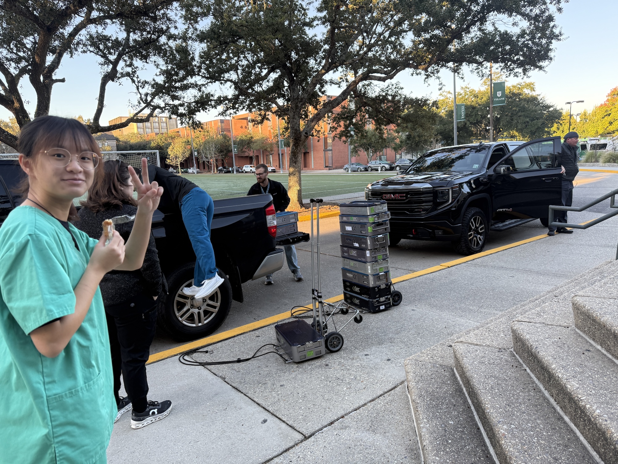 Photograph of a smiling nurse in green scrubs holding up a peace sign, with a truck and crew outdoors.