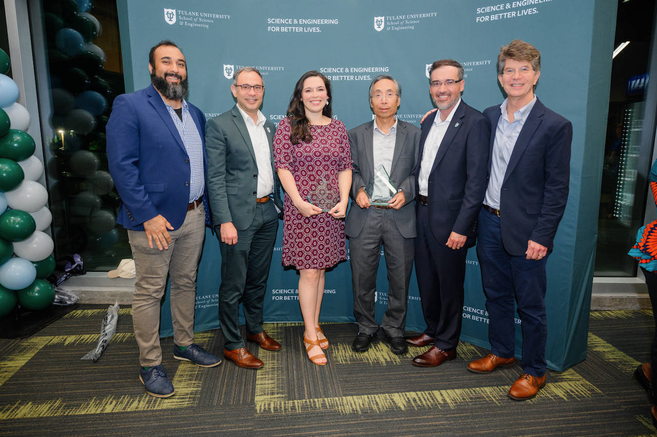 Photograph of six people standing together in front of a blue backdrop, two holding awards.
