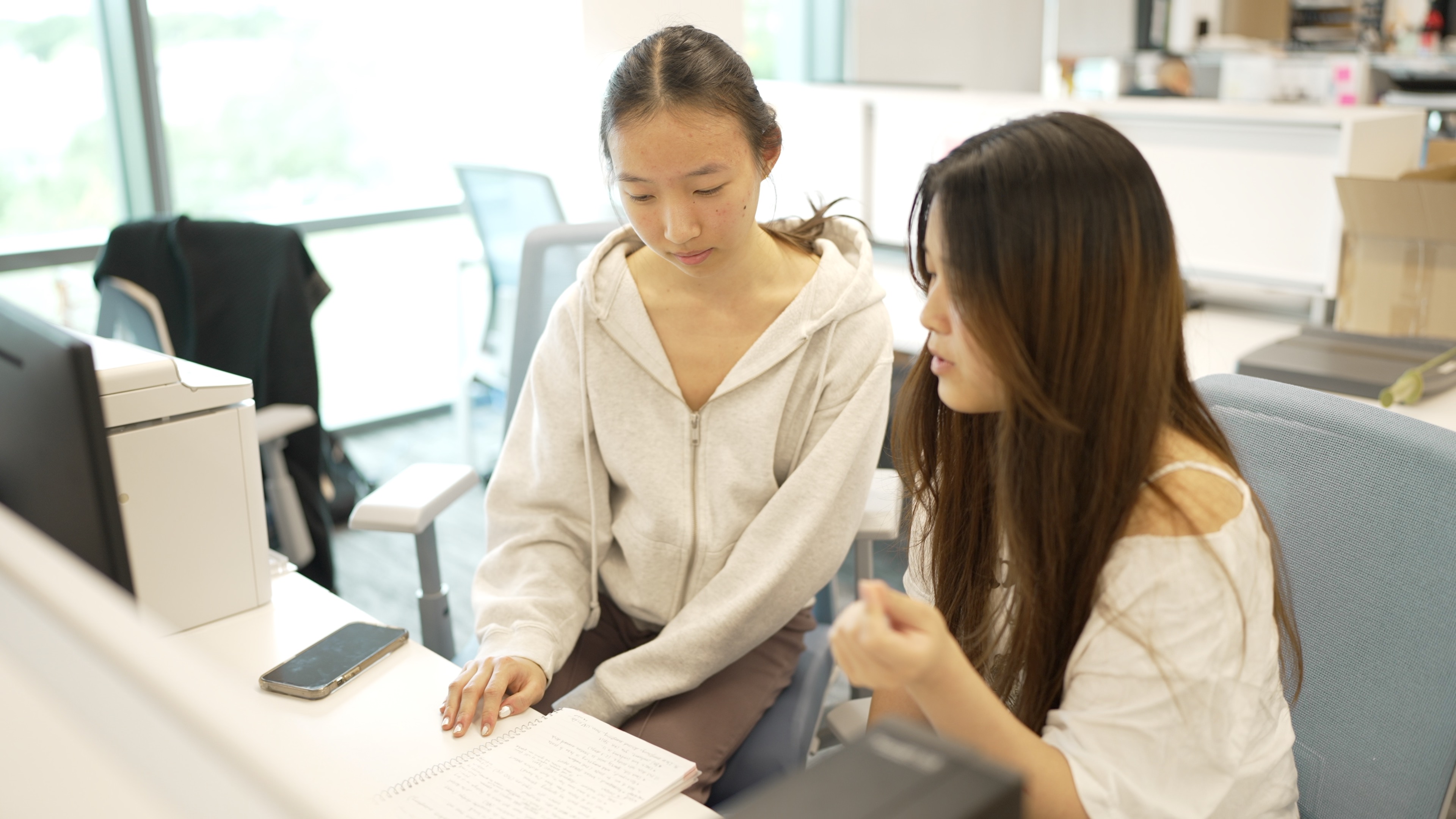 Two young women collaborate at a brightly lit office desk.